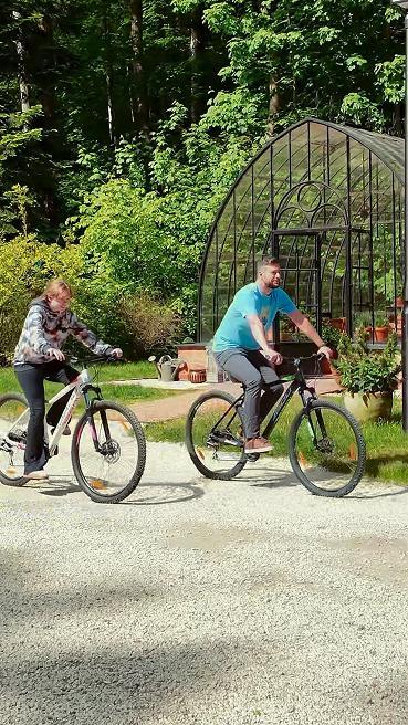 Goście jadący na rowerach obok ogrodu i szklarni przy obiekcie agroturystycznym